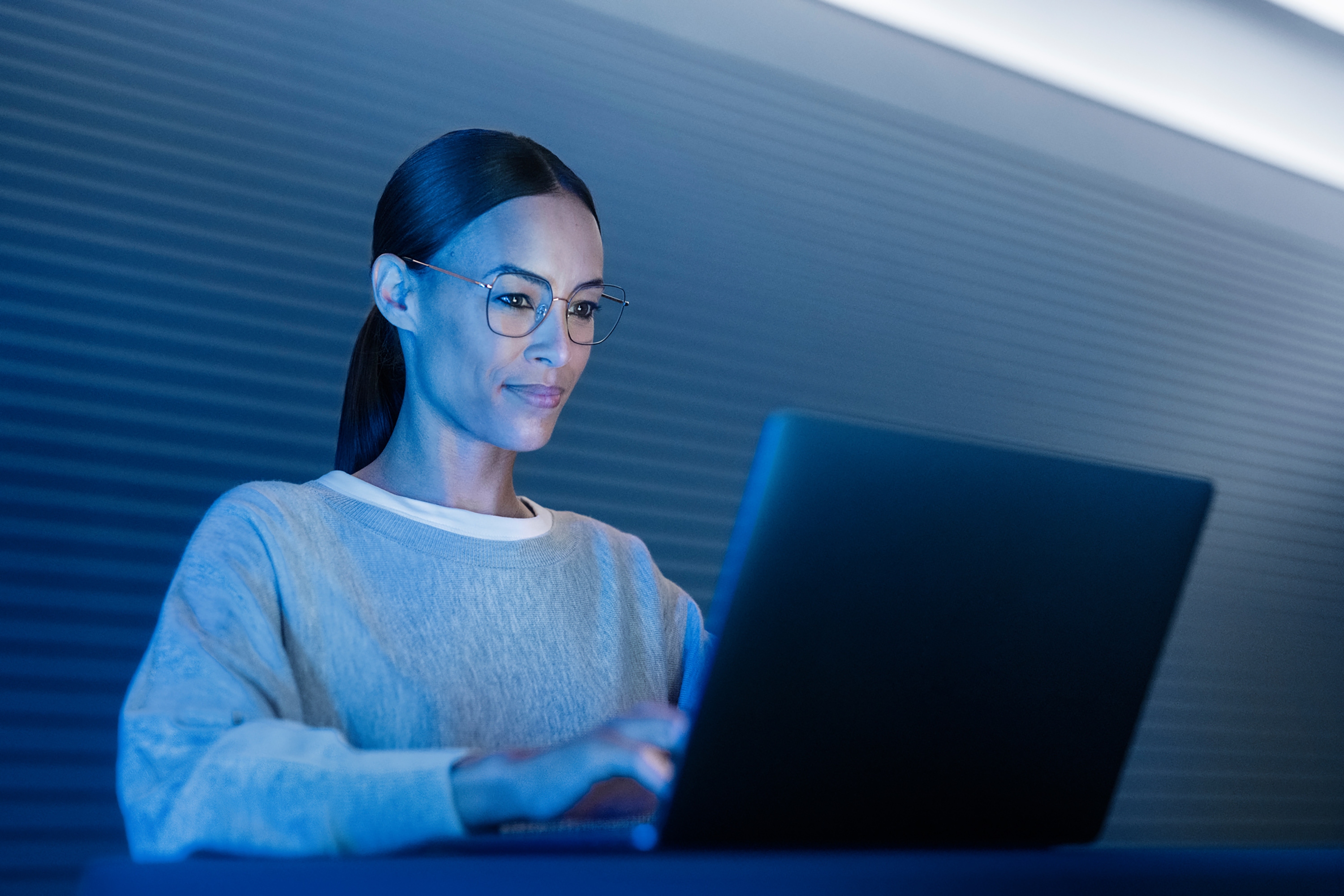 Woman wearing glasses working on a laptop in low light, representing blue light filtering lenses